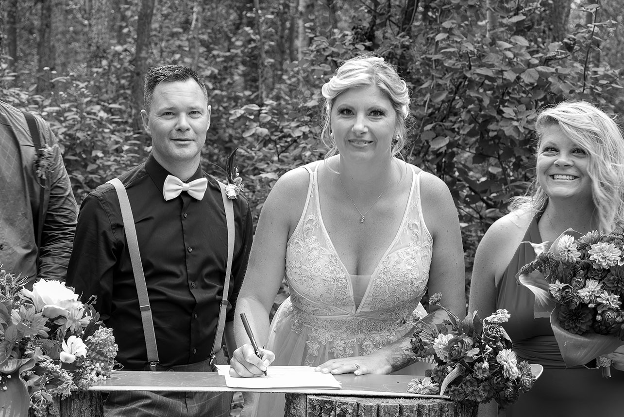 Bride signing wedding registry with groom and bridesmaid smiling beside her